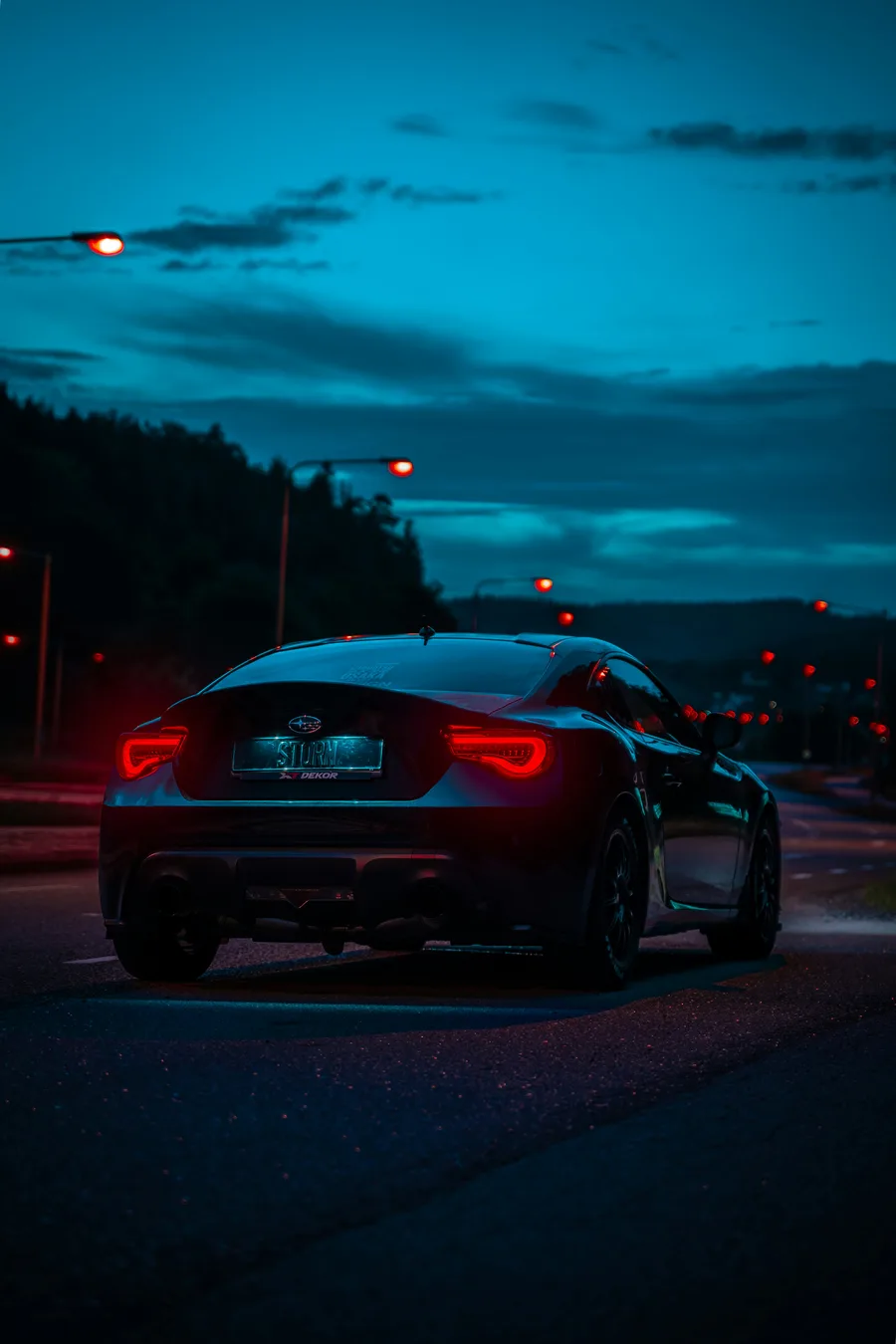 A car with glowing red tail lights on a road at dusk under a teal sky